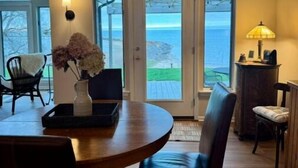 Un comedor con una mesa redonda, sillas, un jarrón con flores y una lámpara. Hay dos puertas que dan a un balcón con vistas al océano.