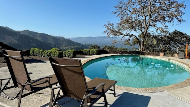 Una piscina con sillas y una mesa, rodeada por un muro de piedra y un árbol, con vistas a montañas al fondo.