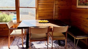 Interior de una cabaña de madera con una mesa de comedor, cuatro sillas, un banco y un ventanal que ofrece vistas de un paisaje montañoso.