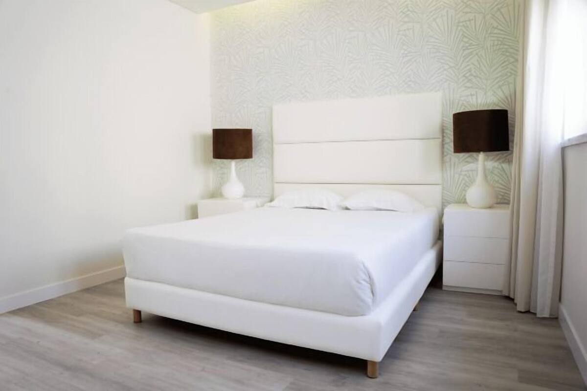 Un dormitorio con una cama blanca, dos mesitas de noche y un papel tapiz estampado.