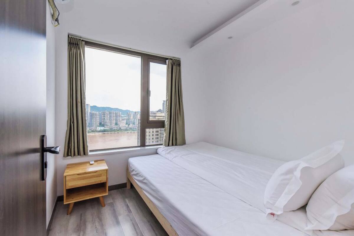 A single-bed room with a wooden nightstand, a window with curtains, and a view of buildings.