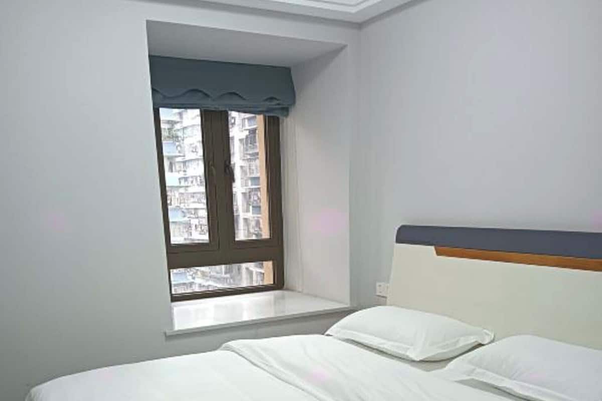 A bedroom with a bed, a window with curtains, and a view of buildings outside.