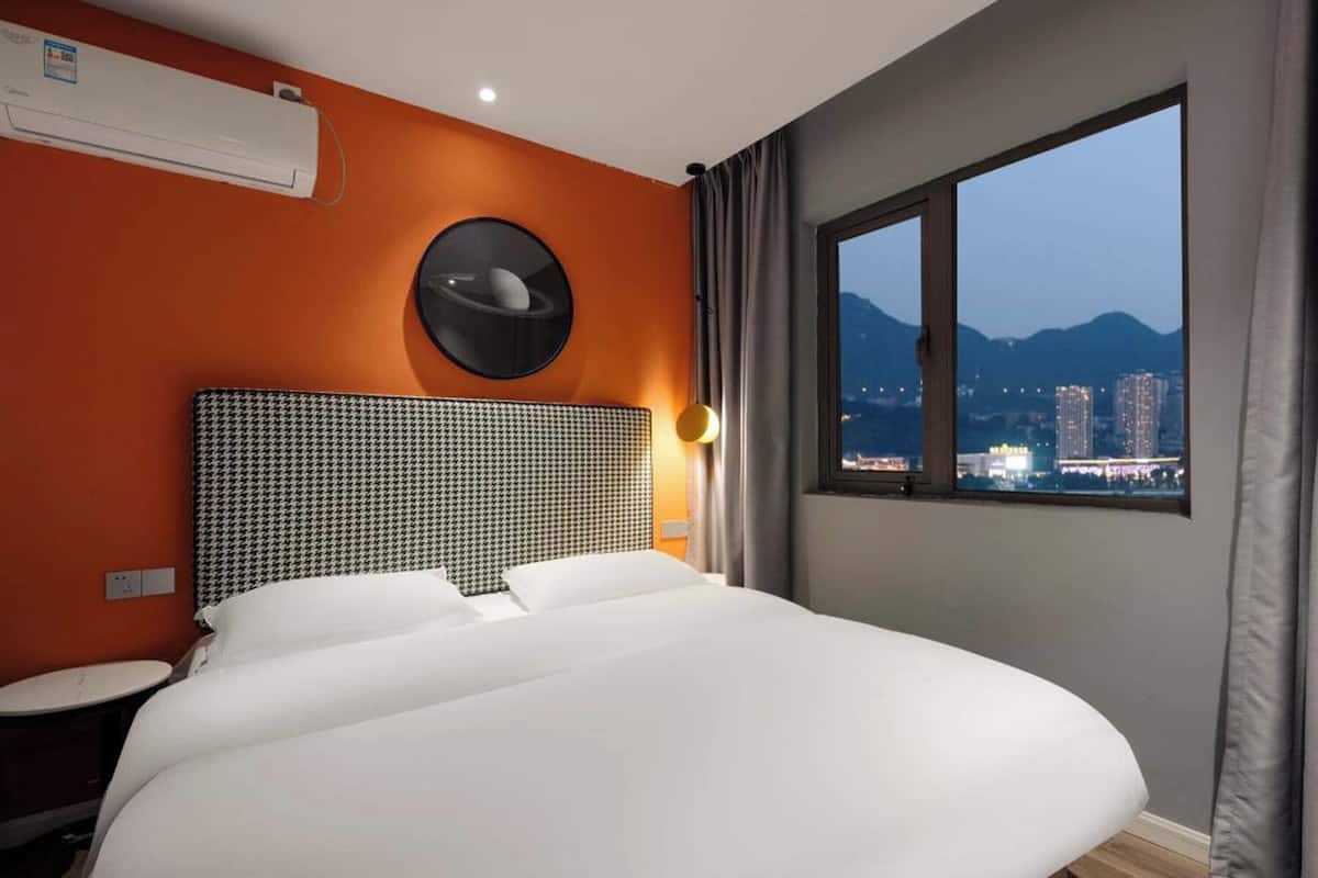 A hotel room with a large bed, an air conditioning unit, a round mirror, and a window with a cityscape view.
