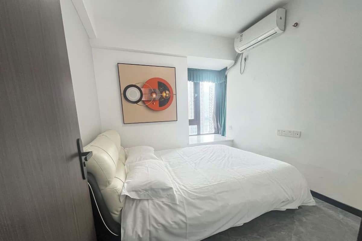 A bedroom with a bed, a wall-mounted air conditioner, a window with curtains, and a framed artwork above the bed.