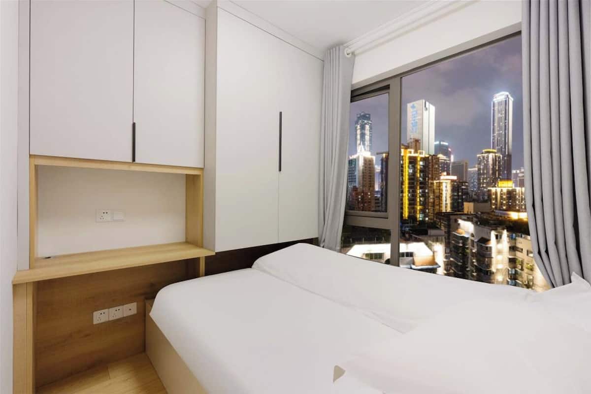 A hotel room with a bed, a wooden nightstand, a white wardrobe, and a large window offering a city view at night.