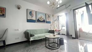 Ein modernes Wohnzimmer mit einem grünen Sofa, einem runden Couchtisch und einem Hängekorbstuhl mit Pflanzen. An der Wand hängen drei gerahmte Kunstwerke und eine Deckenleuchte.