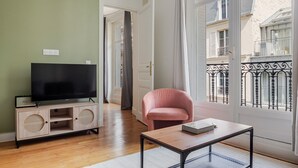 A room with a television on a wooden stand, a pink chair, a small table with a book, and large windows with a view of a balcony. - Blueground | Porte Dauphine, balcony, nr Bois de Boulogne (Paris)