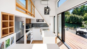 A modern kitchen with a built-in refrigerator, wooden shelving, and a dining area with a view of the garden. - Mountain View Retreat Near Wanaka - Cosy Tiny Home Cottage (Hāwea Flat)