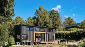 A modern, compact house with a wooden deck, large windows, and a small outdoor seating area. - Mountain View Retreat Near Wanaka - Cosy Tiny Home Cottage (Hāwea Flat)