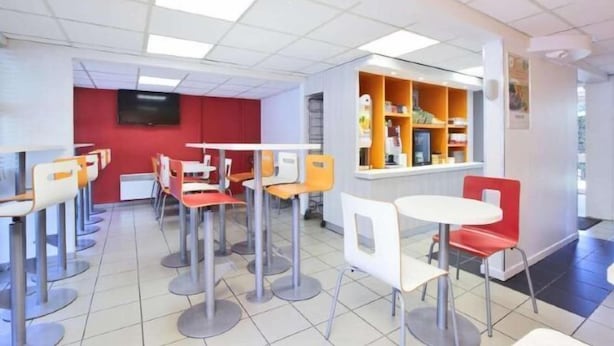 Une cafétéria moderne dotée de nombreuses tables et chaises, d’un mur d’accent rouge et d’une fenêtre avec des étagères.