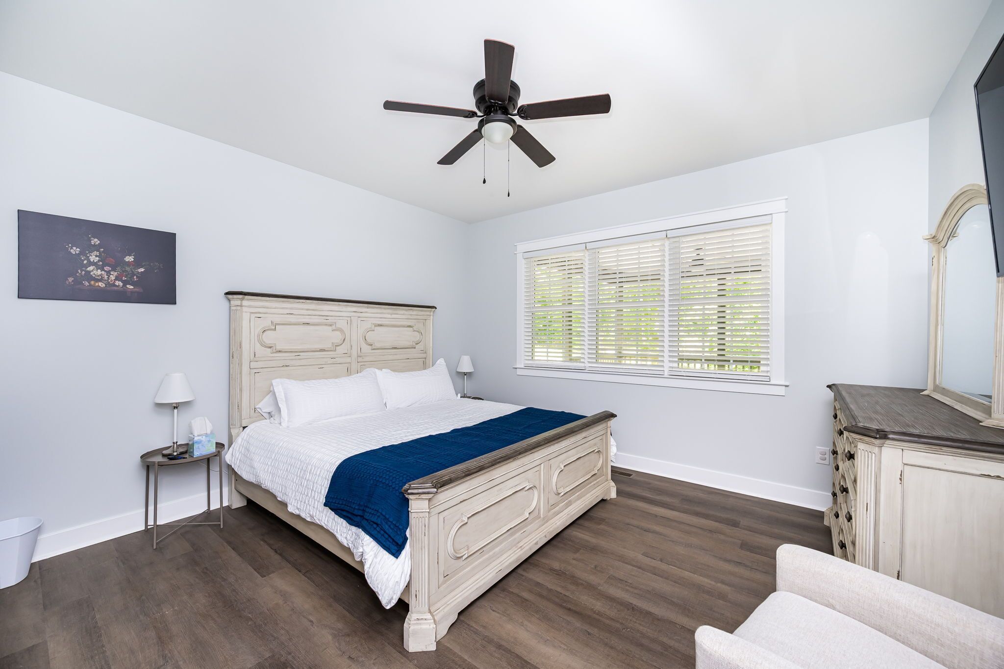 A bedroom with a bed, a ceiling fan, a window with blinds, a nightstand, a lamp, a dresser, and a mirror.