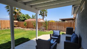 Un patio coperto con mobili in vimini, pavimento in cemento e vista su un giardino privato con recinzione in legno e alberi.