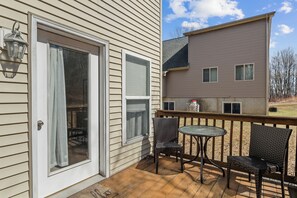 A wooden deck with two wicker chairs and a small table, a sliding glass door, and a view of a two-story house with a brown exterior and multiple windows.