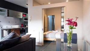 Eine moderne Wohnung mit Küche, einem Essbereich mit Glastisch und einem Schlafzimmer. Ein Badezimmer ist durch eine offene Tür sichtbar.
