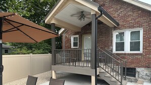 Une terrasse couverte avec une table et des chaises, une maison en briques avec une fenêtre et un escalier menant à l’entrée.