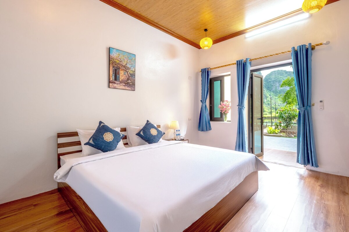 A bedroom with a wooden bed, white bedding, a painting on the wall, and a view through a glass door.