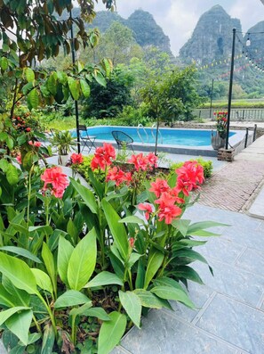 A pool area with a view of mountains, surrounded by lush greenery and red flowers. - Tràng An Sunny River Homestay (Hoa Lu)