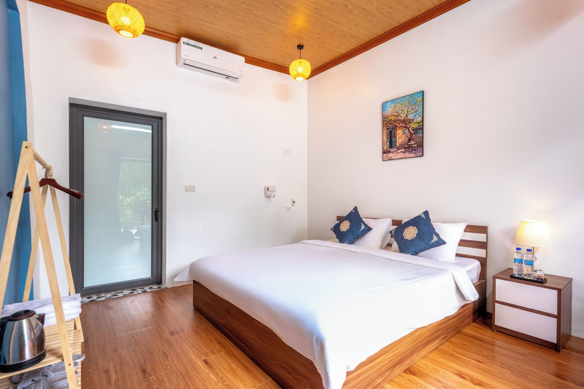 Un dormitorio con cama de madera, ropa de cama blanca, un cuadro en la pared y una mesita de noche con lámpara.
