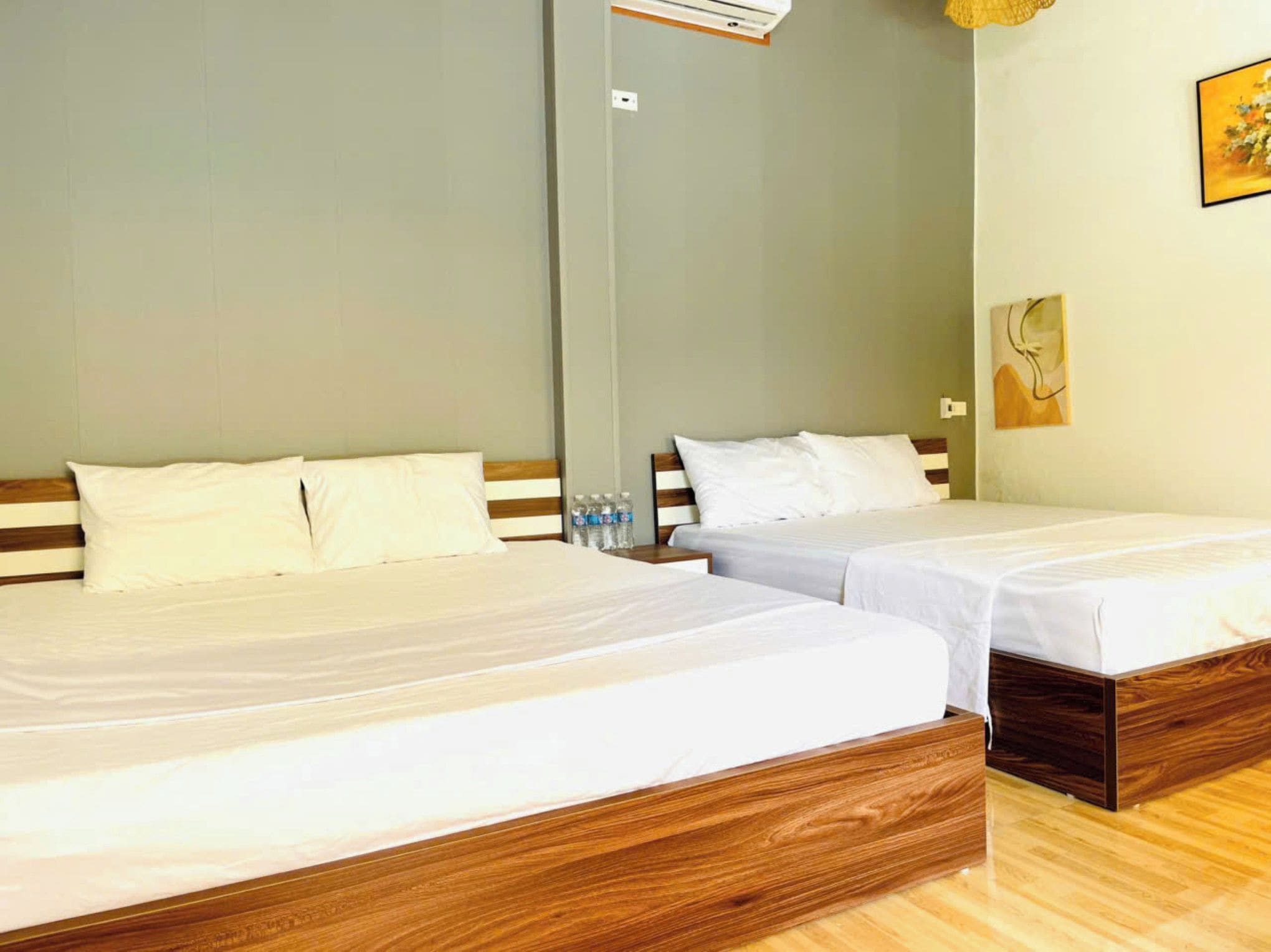 Une chambre d’hôtel avec deux lits simples, des têtes de lit en bois, du linge de lit blanc, un plancher en bois, un climatiseur fixé au mur et une petite table avec des bouteilles d’eau.