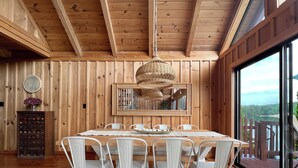 Een eetzaal met houten lambrisering, een grote tafel en witte stoelen, een hanglamp en uitzicht op een meer door grote ramen.