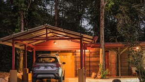 Un automóvil estacionado bajo un garaje cubierto con estructura de madera y un pequeño jardín con rocas y plantas.