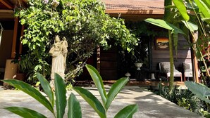 Un patio con una estatua de piedra, abundante follaje y un edificio de madera con techo de tejas.