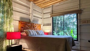 Un dormitorio con techo de madera, una cama con cabecera, una ventana con vistas a follaje y una lámpara roja al lado de la cama.