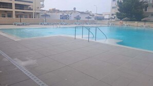 Ein Schwimmbecken mit Fliesenrand, Liegestühlen und einem Gebäude mit Balkonen im Hintergrund.