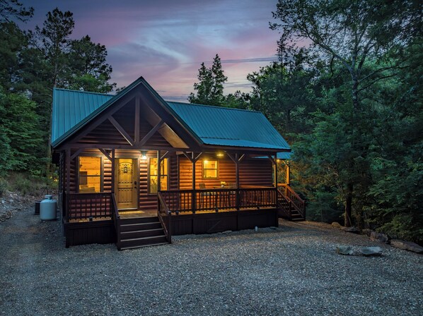 Situated in a wooded area, this cabin features a rustic gabled roof and a welcoming porch with a decorative front door.