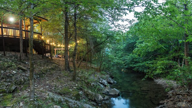 Eine rustikale Holzhütte mit einer Holzdiele und Lichterketten, umgeben von dichtem Wald und einem klaren Bach.