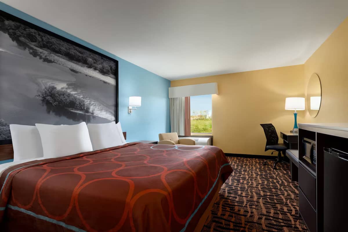 Habitación de hotel con una cama grande, un escritorio con computadora, una silla y vista al exterior a través de una ventana.