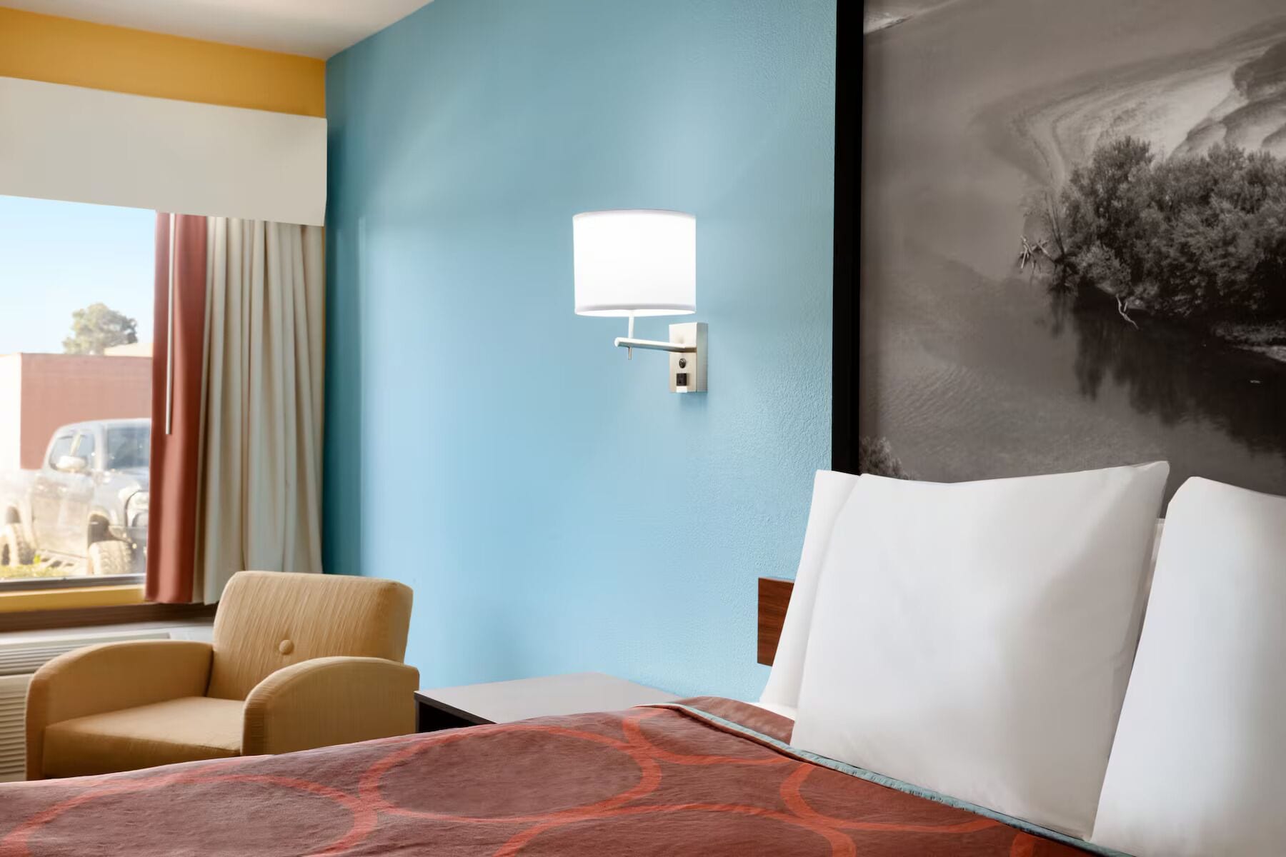A hotel room with a bed, a chair, a wall-mounted lamp, and a large painting.