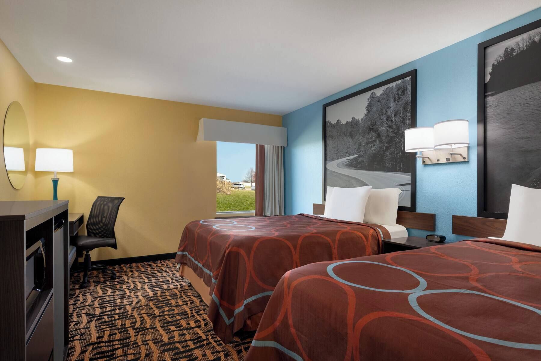 A hotel room with two beds, a desk, a chair, a window with a view, and wall-mounted lamps.