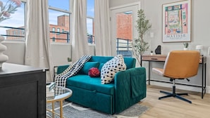 A modern living room with a teal sofa, a small round table, a desk with a chair, and a large window with curtains. - Cozy 1BR|1BA Retreat in Philly (Philadelphia)