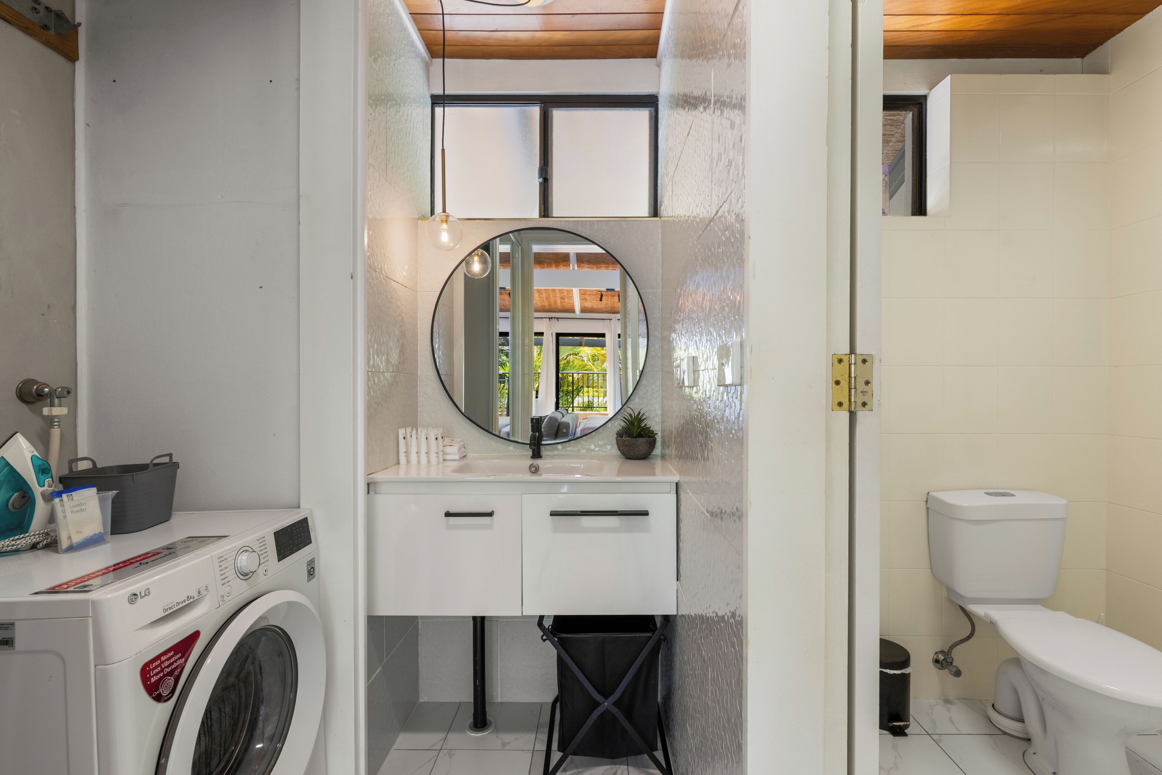 Une petite salle de bain équipée d’un lave-linge, d’un lavabo avec un miroir et de toilettes.