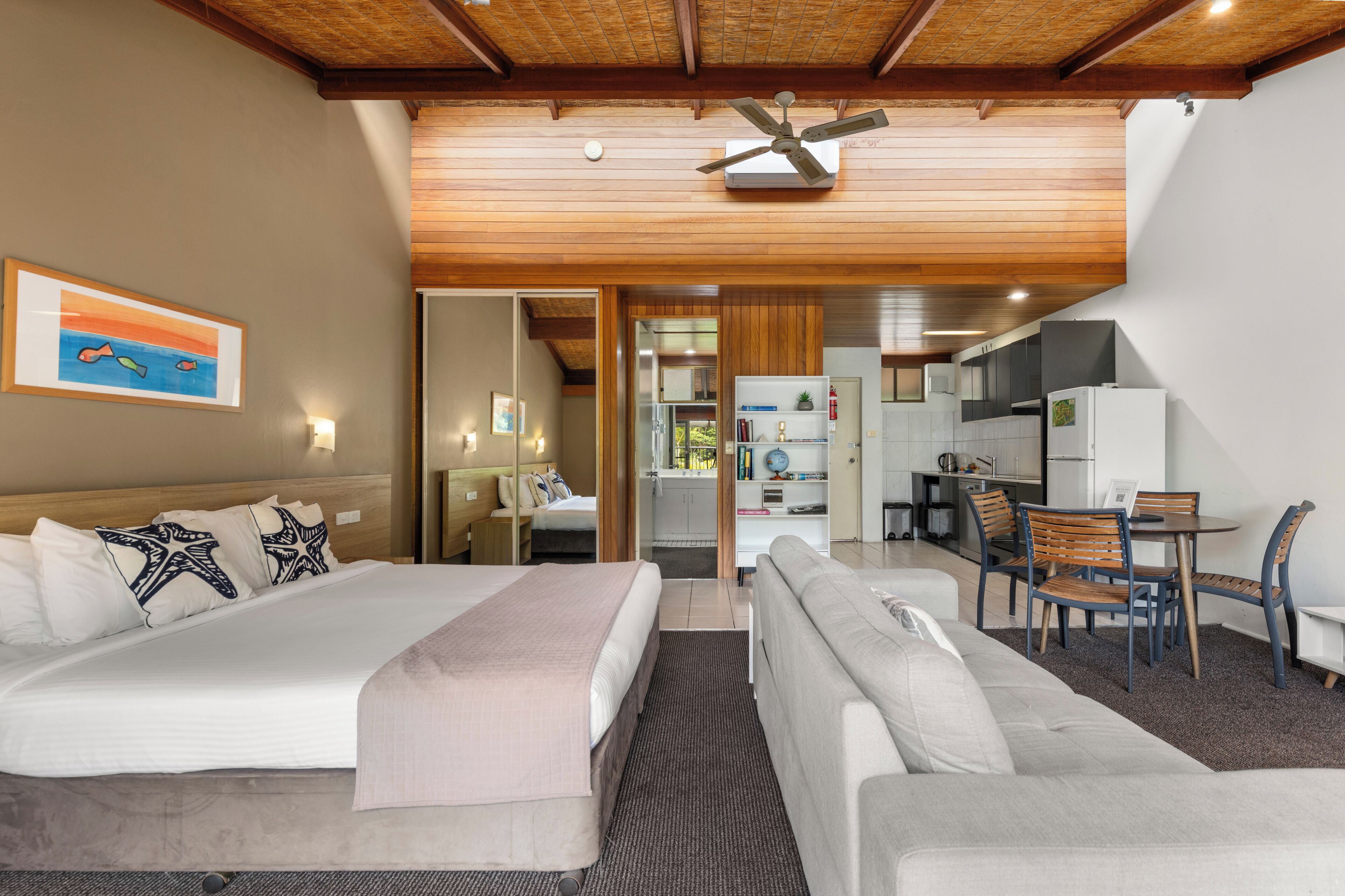 A hotel room with a bed, a sofa, a dining table with chairs, a kitchen area, and a ceiling fan.