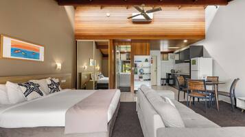 A hotel room with a bed, a sofa, a dining table with chairs, a kitchen area, and a ceiling fan.