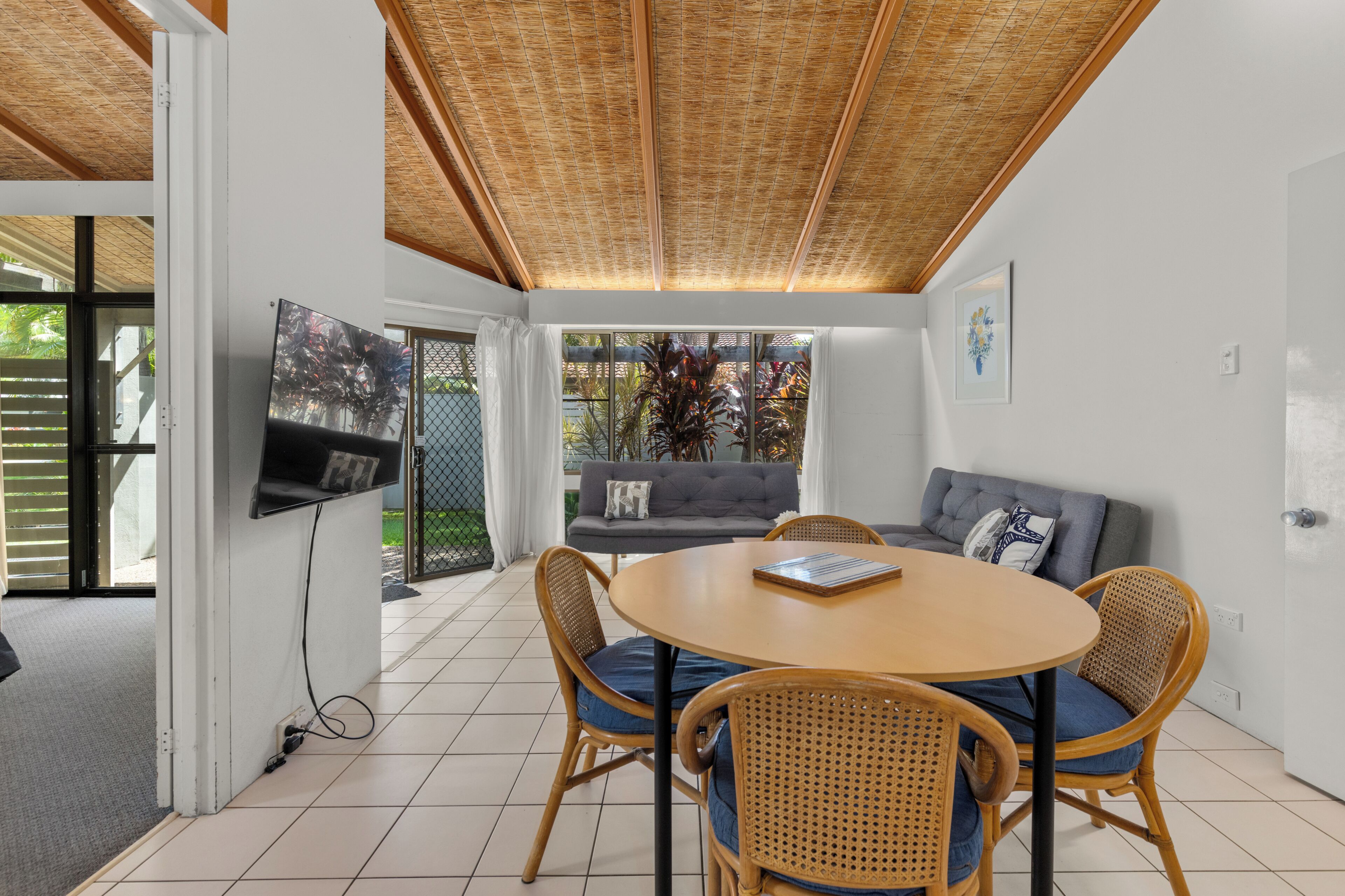 A modern living room with a round wooden table, wicker chairs, a grey sofa, and a TV mounted on the wall.