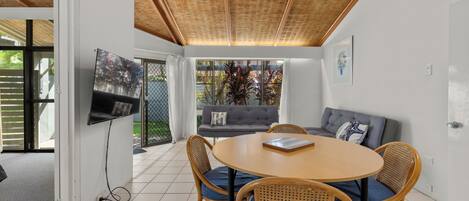 A modern living room with a round wooden table, wicker chairs, a grey sofa, and a TV mounted on the wall.