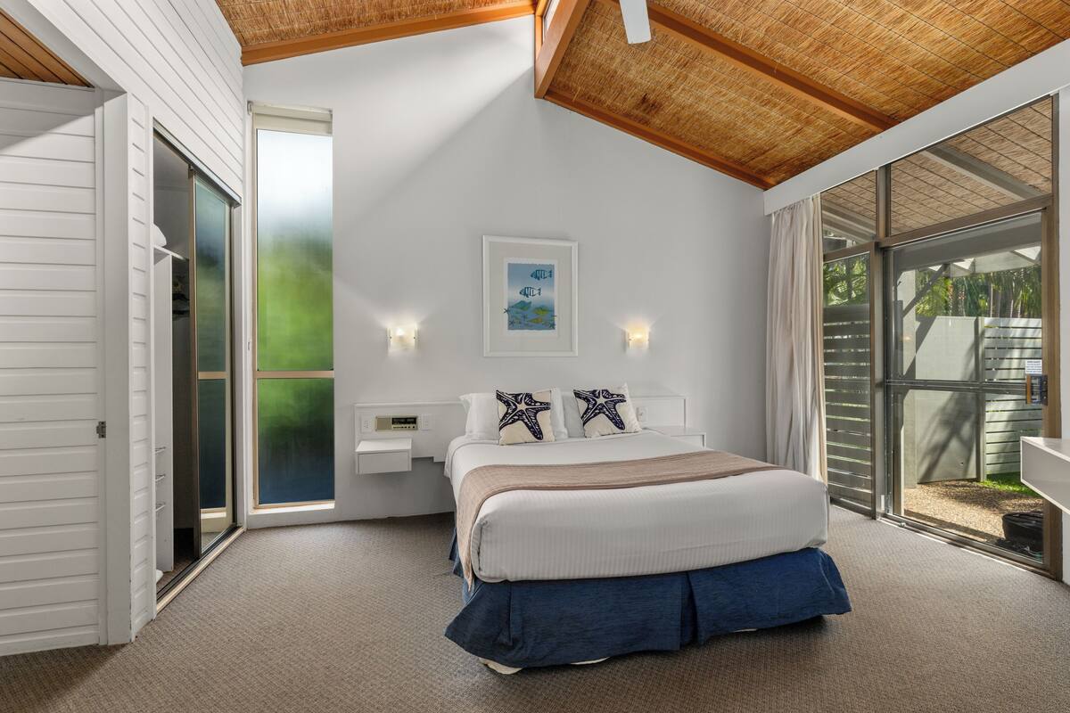 Ein Schlafzimmer mit einem Bett, einem Nachttisch, einer Wandleuchte, einem gerahmten Bild und einer Glasbodentür, die zu einem Außenbereich führt.