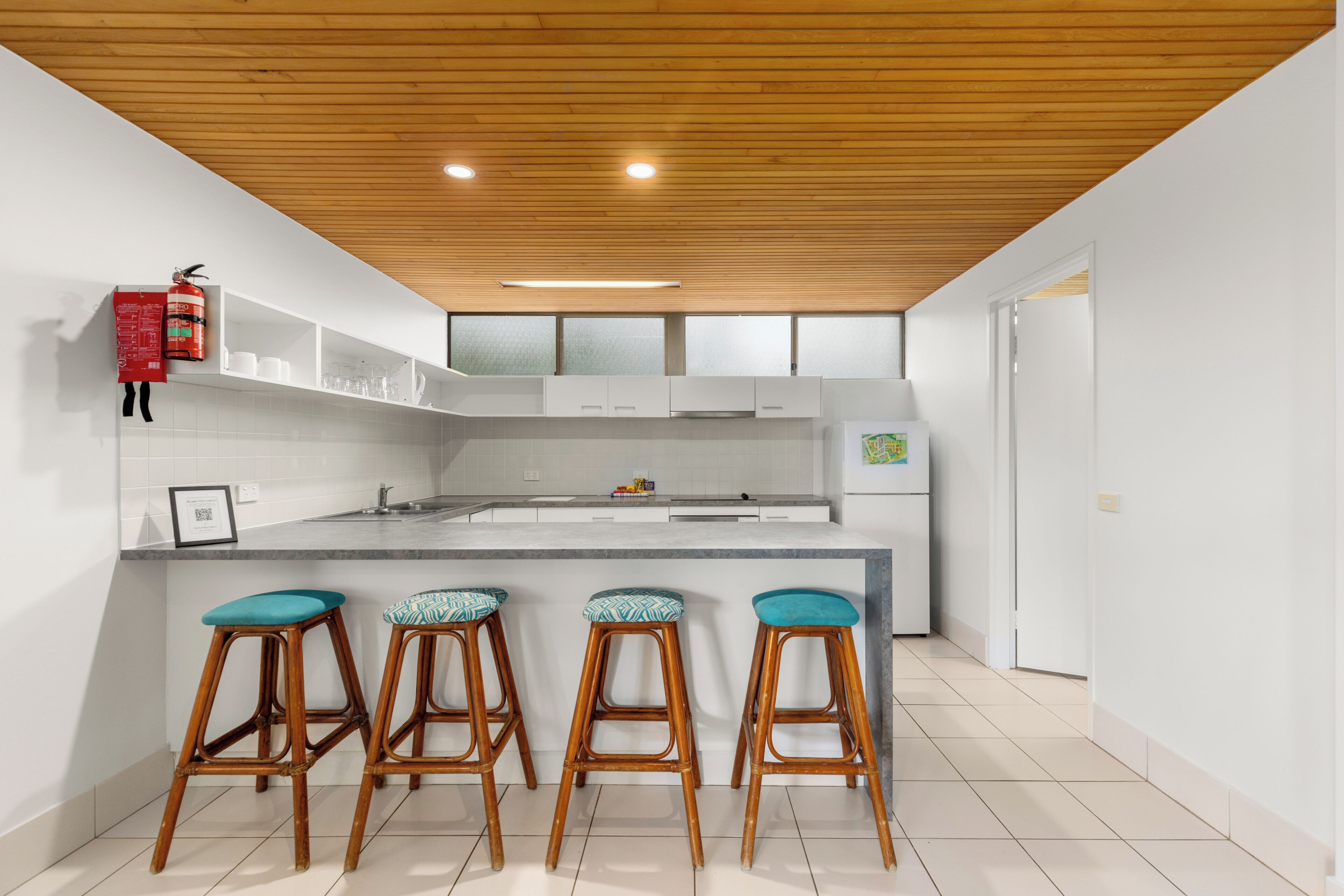A modern kitchen with a wooden ceiling, white cabinets, a central island with stools, and a fire extinguisher on the wall.