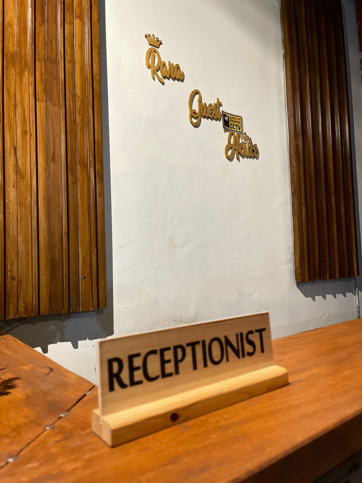A reception desk with a sign that reads "RECEPTIONIST" and decorative wall signs that read "Rara Guest Area CCTV."