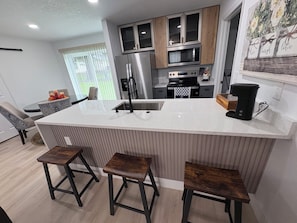 A modern kitchen with a central island, stainless steel appliances, and wooden bar stools. - Cozy 3BR, 2 BAT Near the World Equestrian Center. (Ocala)