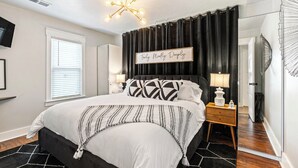 A bedroom with a bed, bedside tables, a chandelier, a black curtain, a sign on the wall, and a door.