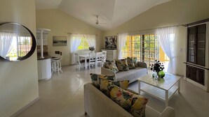 A spacious living room with a sofa, coffee table, dining table, and chairs. There is a large window with curtains, a ceiling fan, and a view of the outdoors.