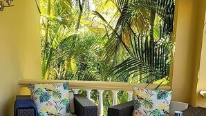 A sunlit room with a sofa set against a yellow wall, a small table with a vase of flowers, and a view of tropical foliage through a window.