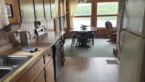 Une cuisine avec des meubles en bois, une table à manger avec des chaises, un réfrigérateur et une vue sur un lac, visible à travers de grandes fenêtres.