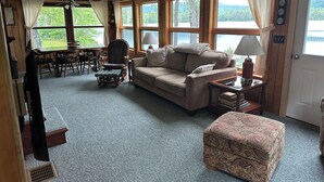 Une pièce chaleureuse avec un canapé, un pouf à motifs, une table à manger avec des chaises et de grandes fenêtres donnant sur l’extérieur.