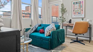 Een moderne woonkamer met een blauwgroene bank, een houten bureau en een uitzicht op gebouwen door grote ramen.