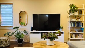 A living room with a flat-screen TV, a round wooden coffee table, a sofa, a bookshelf, and several potted plants. - Charming and Spacious 2B + 2.5 Bath Townhome (Pasadena)